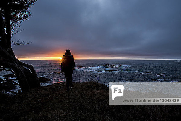 Silhouette teenage girl looking at view by sea against cloudy sky during sunset