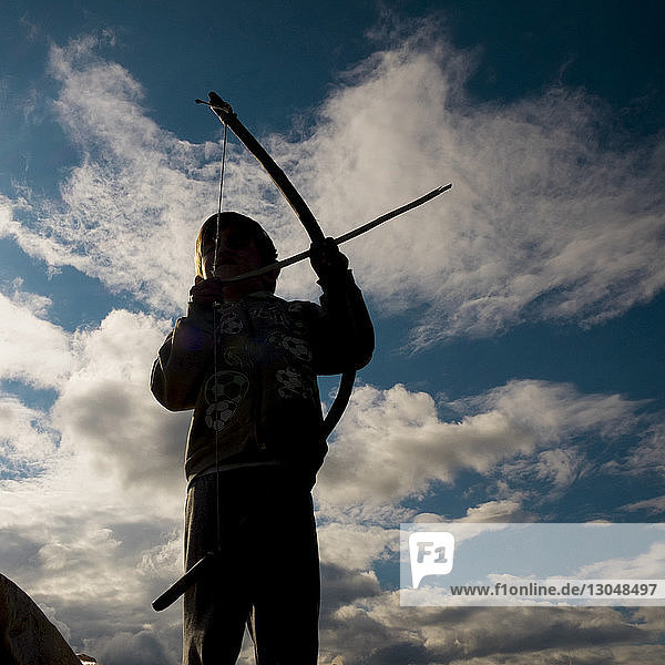 Low angle view of boy holding bow and arrow against cloudy sky