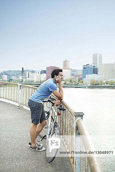 Männlicher Sportler  der ein Mobiltelefon benutzt  während er mit dem Fahrrad auf einer Brücke am Fluss steht