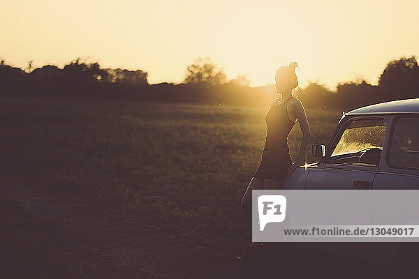 Frau steht bei Sonnenuntergang am Auto auf dem Feld gegen den Himmel