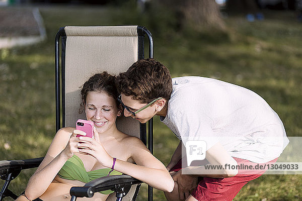 Bruder sieht Schwester mit Smartphone an  während er auf einem Stuhl im Rasen sitzt
