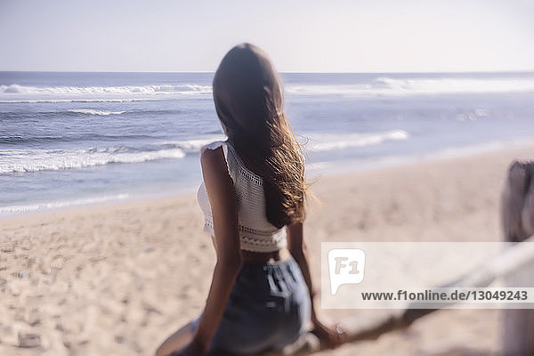 Frau betrachtet Aussicht  während sie auf einem Holzzaun am Strand sitzt