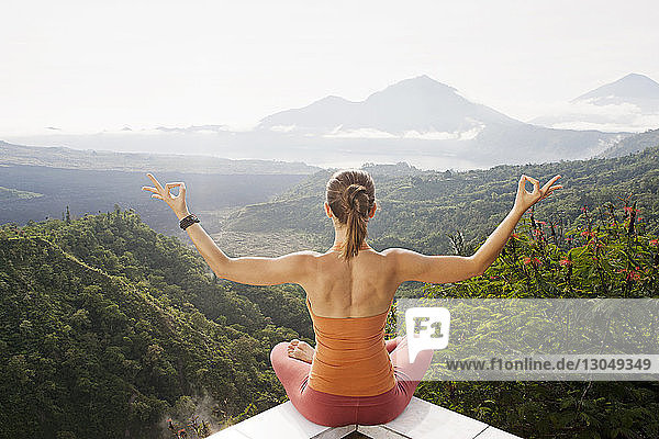 Rear view of woman meditating while sitting on retaining wall by mountains