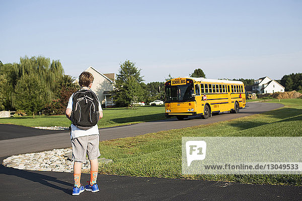 Rückansicht eines Jungen mit Rucksack  der auf den Schulbus wartet  gegen den klaren Himmel auf dem Rasen
