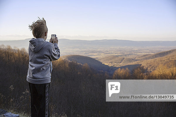 Rückansicht eines Jungen  der mit einem Mobiltelefon fotografiert  während er auf einem Berg vor klarem Himmel steht