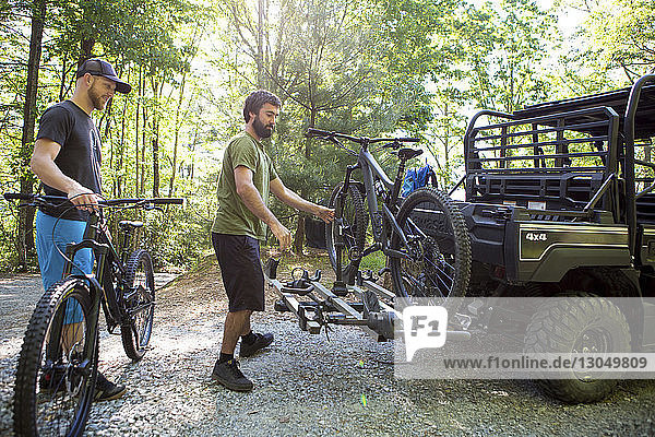 Man looking at friend putting mountain bike on off-road vehicle at forest