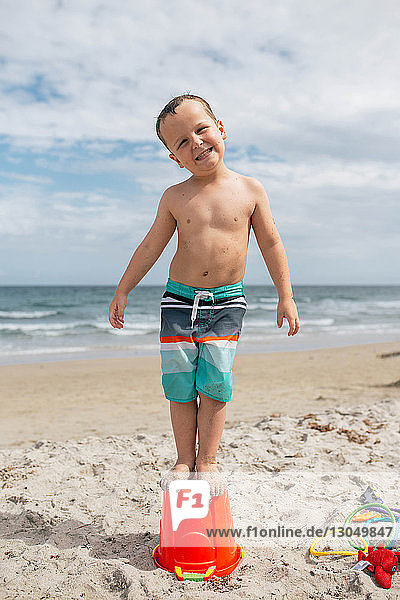 Portrait of playful boy standing on bucket at beach against cloudy sky
