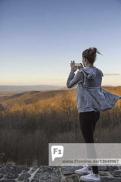 Rückansicht einer Teenagerin  die mit einem Mobiltelefon fotografiert  während sie auf einem Berg vor klarem Himmel steht