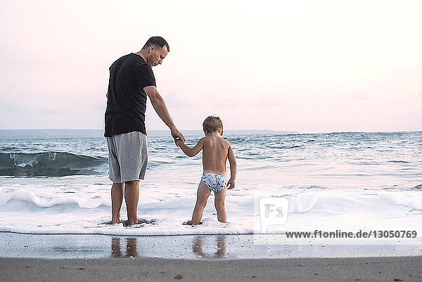 Side view of father with shirtless son standing at beach against clear sky during sunset