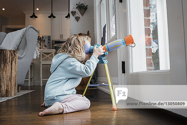Side view of girl looking through telescope while kneeling by door at home