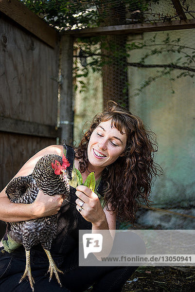 Fröhliche Frau füttert kauernd in der Geflügelfarm eine Henne mit Blättern