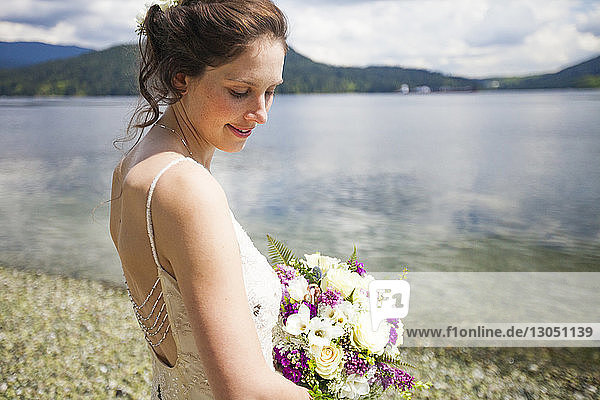 Seitenansicht der am Seeufer stehenden lächelnden Braut mit Blumenstrauss