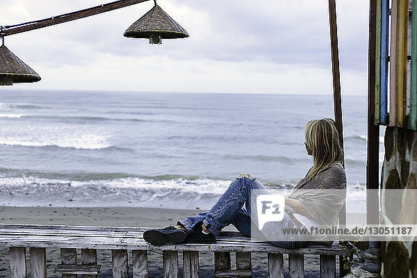 Seitenansicht einer Frau  die auf einem Holzgeländer am Strand vor bewölktem Himmel sitzt