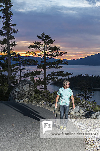 Mann hält Skateboard und schaut weg  während er auf einem Fußweg am See geht