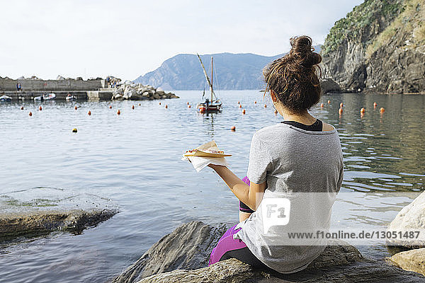 Rückansicht einer Frau  die auf einem Felsen am Meer sitzend Pizza isst