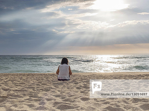 Rückansicht einer Frau  die bei Sonnenuntergang auf Sand am Strand vor bewölktem Himmel sitzt