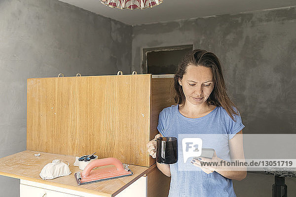 Frau mit Getränk beim Telefonieren  während sie während der Renovierung zu Hause an den Möbeln steht