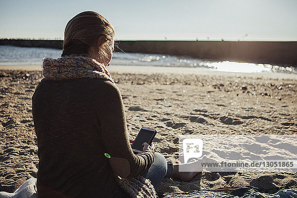 Rückansicht einer Frau  die ein Smartphone benutzt  während sie am Strand im Sand sitzt