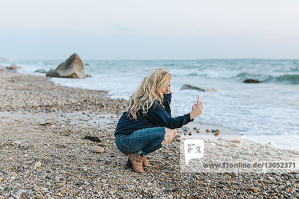 Junge Frau kauert am Strand und schaut auf ihr Smartphone, Menemsha, Martha's Vineyard, Massachusetts, USA