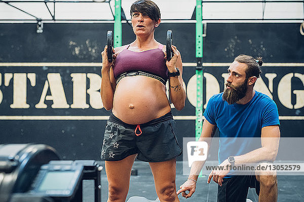 Trainer guiding pregnant woman using weights in gym