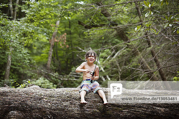 Smiling boy fishing while sitting on fallen tree trunk in forest