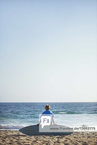 Rear view of boy sitting on surfboard at beach against clear sky