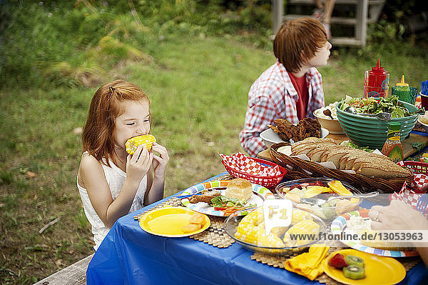 Mädchen isst Zuckermais  während sie mit ihrem Bruder am Picknicktisch sitzt