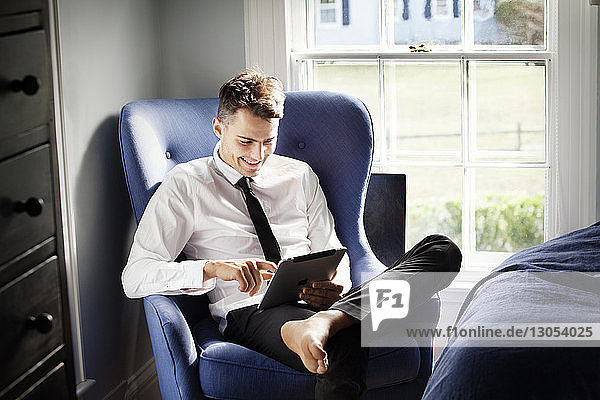 Glücklicher Mann benutzt Tablet-Computer  während er sich zu Hause auf einem Sessel entspannt