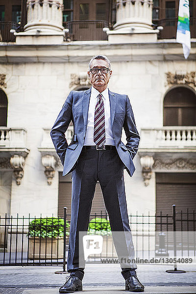 Portrait of senior businessman with hands in pockets standing against building