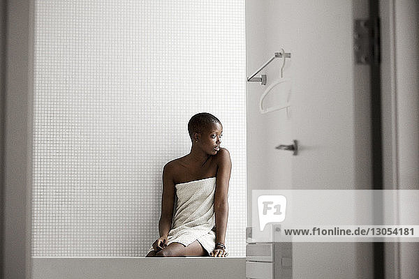 In ein Handtuch gewickelte Frau schaut weg  während sie zu Hause auf der Badewanne sitzt