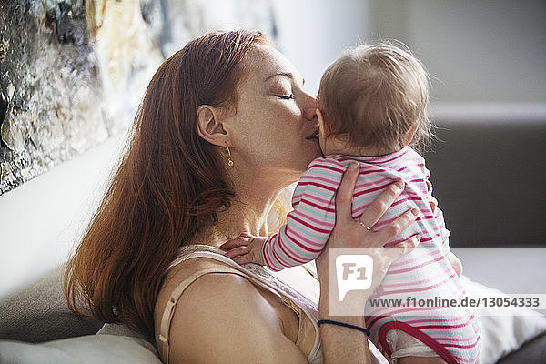 Mother kissing baby girl while sitting on sofa at home
