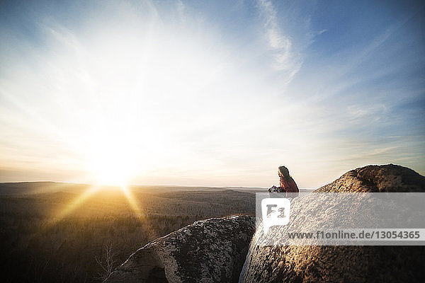 Side view of woman sitting on rocks at cliff against sky during sunset