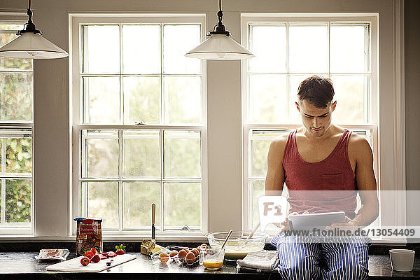 Mann benutzt Tablet-Computer für die Rezeptsuche  während er zu Hause auf der Küchentheke sitzt