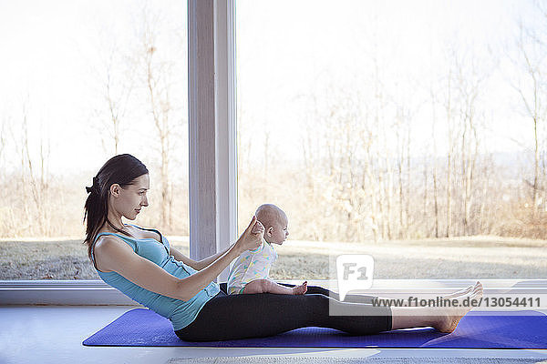 Side view of woman practicing yoga while holding baby girl by window