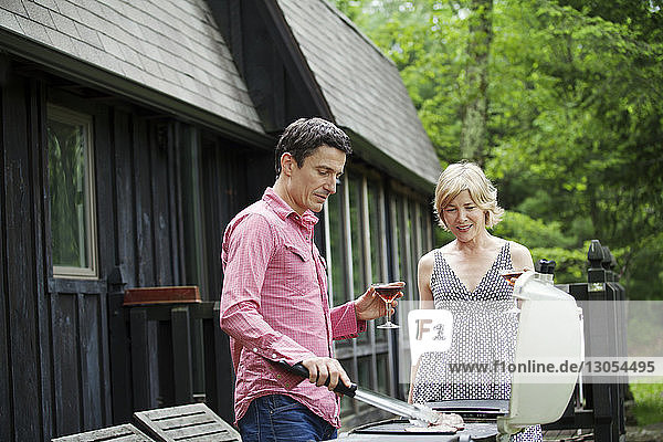 Man preparing food on barbecue grill while standing with wife by cottage