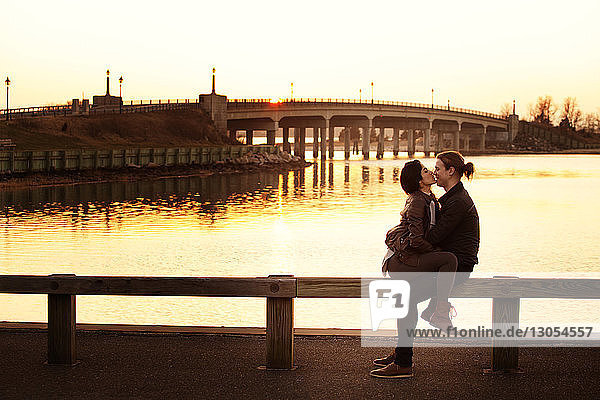 Romantic couple sitting on railing against sky during sunset