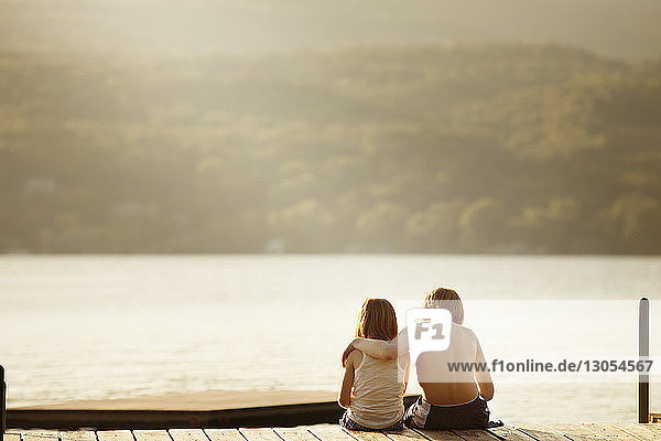 Rückansicht des Bruders  der mit der Schwester auf dem Steg über dem See Arm um Arm sitzt