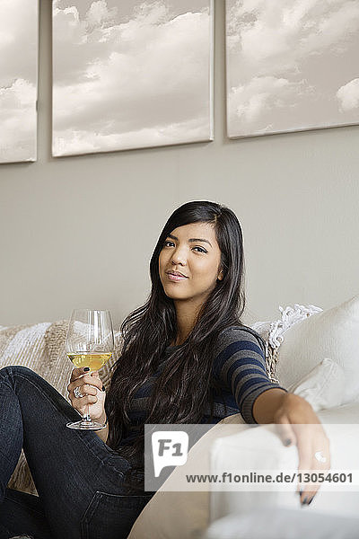 Lächelnde Frau hält Weinglas in der Hand  während sie zu Hause auf dem Sofa sitzt