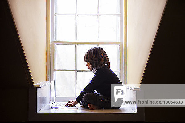 Junge benutzt Laptop-Computer  während er zu Hause am Fenster sitzt