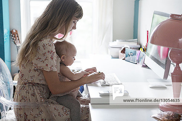 Mutter  die ihr Baby hält  während sie zu Hause einen Desktop-Computer benutzt