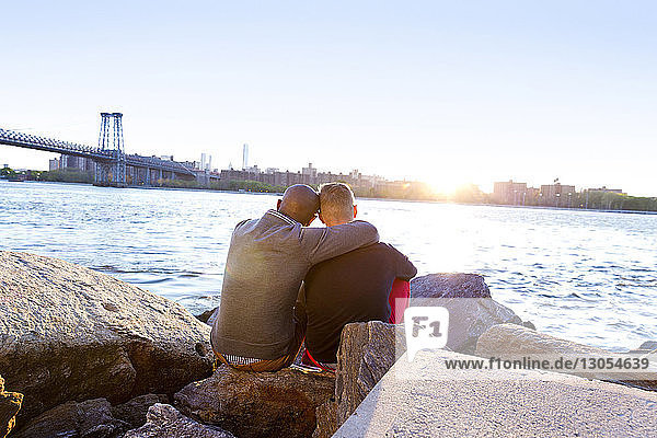Rear view of man sitting arm around with boyfriend on rocks at East River during sunset