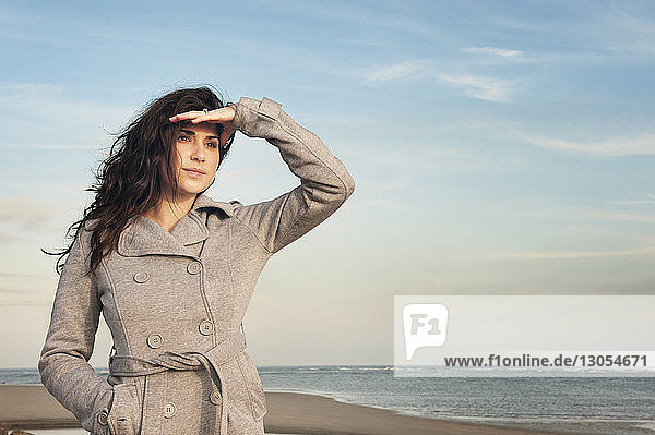 Woman shielding eyes while standing on beach against sky