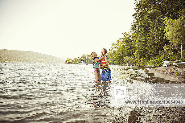 Cheerful siblings embracing while wearing life jackets in lake against clear sky