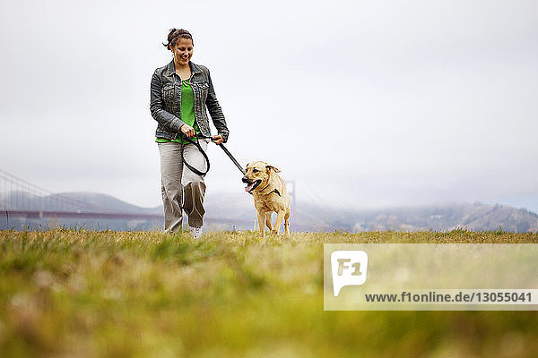 Happy woman walking with dog on grassy field against cloudy sky