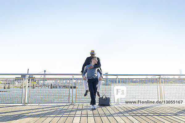 Happy man embracing boyfriend on pier by river against clear sky