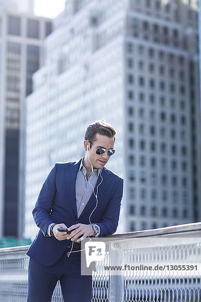 Geschäftsmann hört Musik am Telefon  während er sich in der Stadt an ein Geländer lehnt