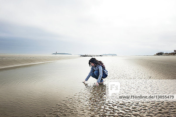 Woman crouching on beach against cloudy sky