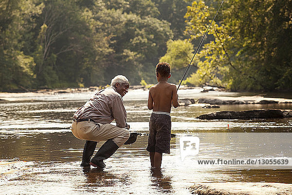 Grandfather crouching while looking at grandson fishing at lake