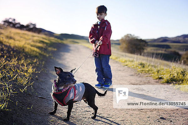 Boy holding pet leash of dog while standing on footpath by field