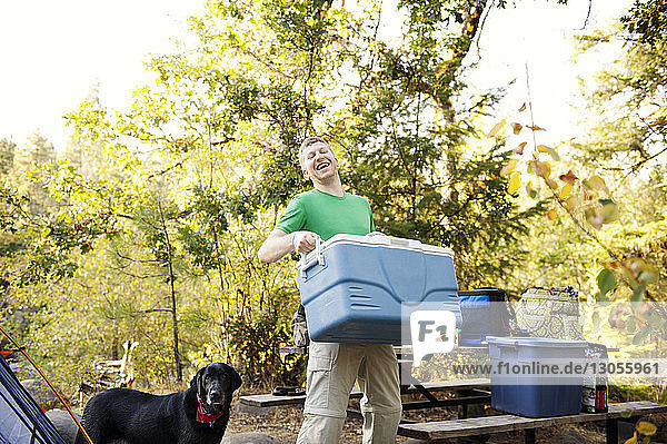 Glücklicher Mann trägt Kühlbox  während er auf dem Campingplatz bei seinem Hund steht